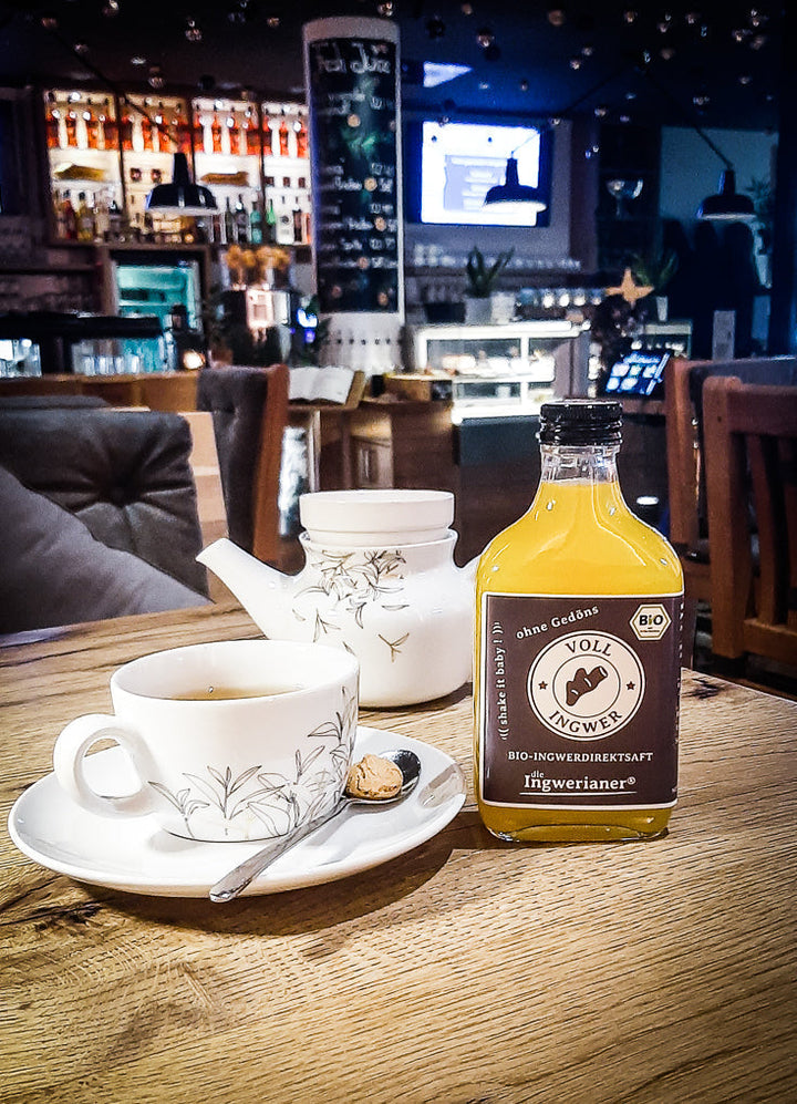 Eine Tasse und eine Kanne Ingwertee mit einer Flasche Ingwerianer®, auf einem Tisch stehend.